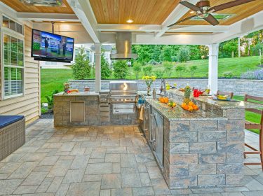 An outdoor kitchen containing a stone bar complete with a grill and large television mounted overhead