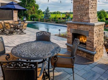 An outfoor fireplace on a stone patio that surrounds a pool with chairs and a table