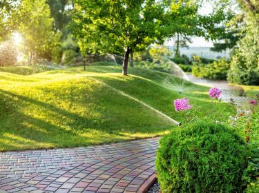 A brick path lined with flowers winding through lush landscaping that is being watered with sprinkers